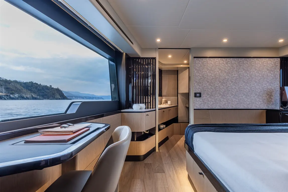 Luxury yacht interior featuring a modern bedroom, desk, and large window overlooking the sea and distant landscape.